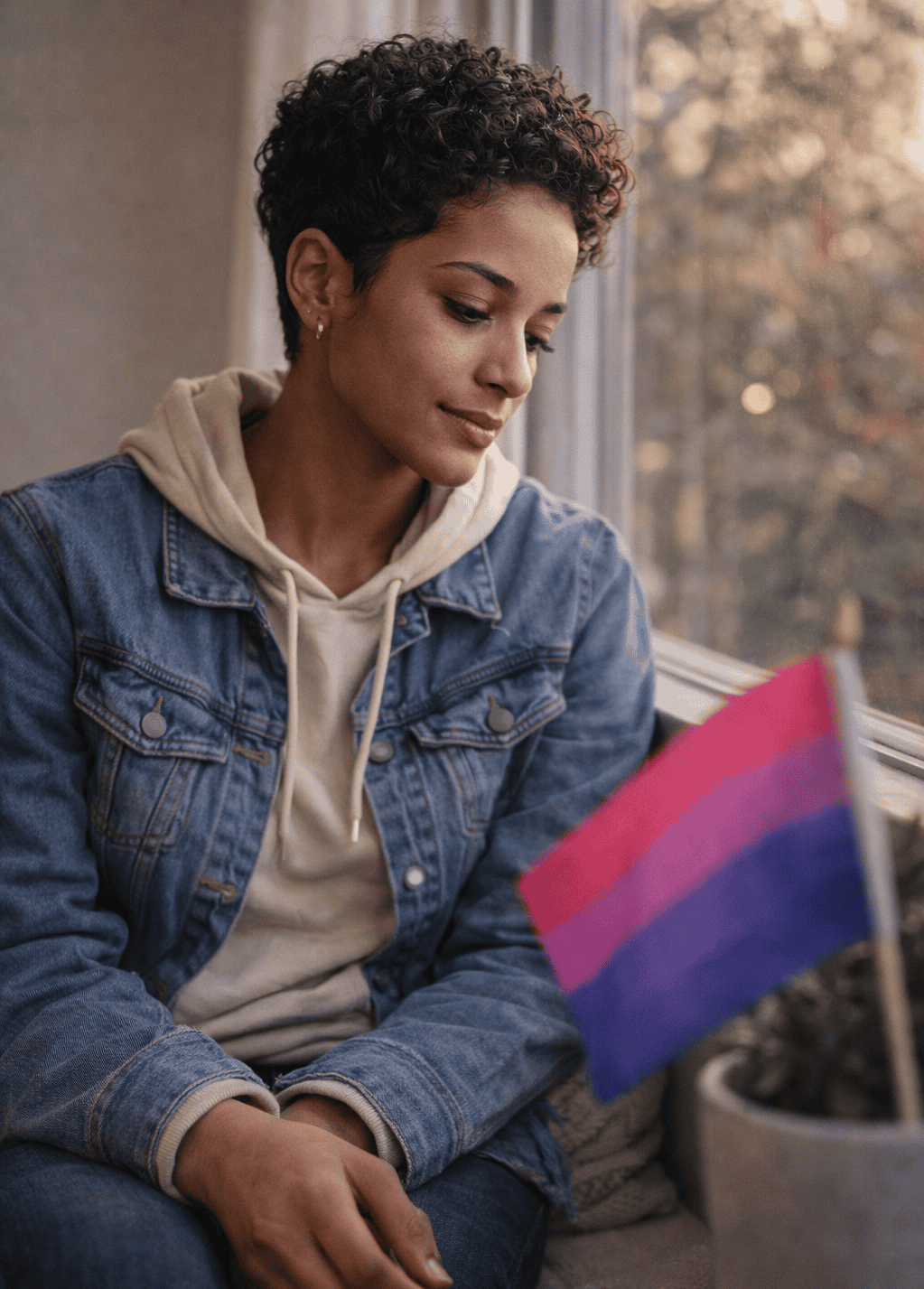 Young person sitting thoughtfully by a window with a bisexual pride flag nearby, representing bisexual mental health and emotional support