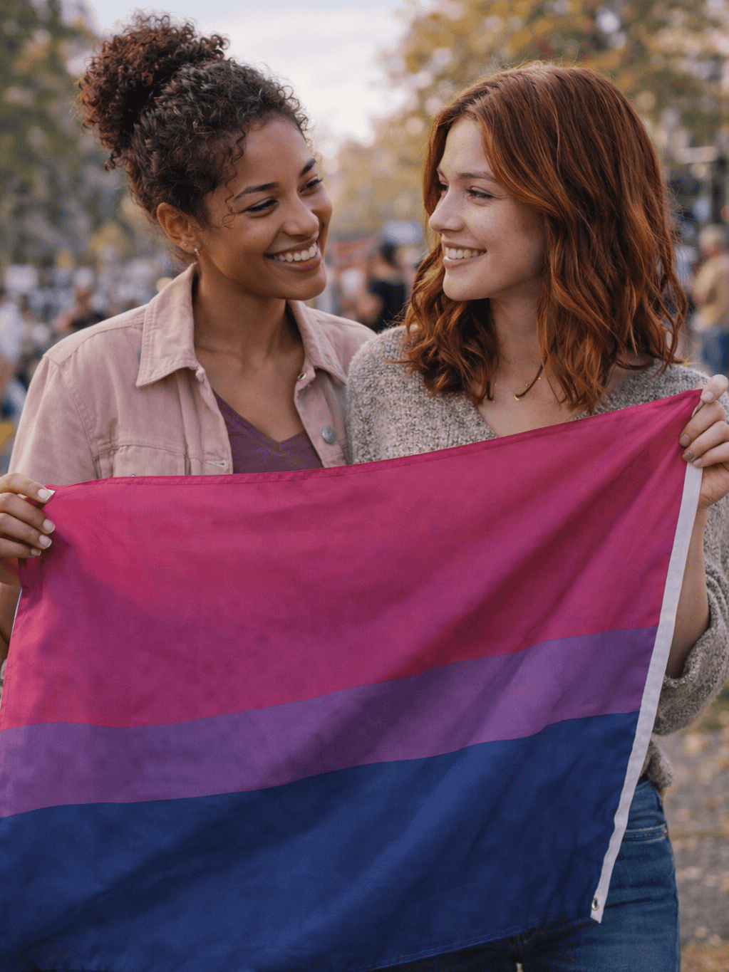 Two women smiling and holding a bisexual pride flag, representing bisexual visibility and identity